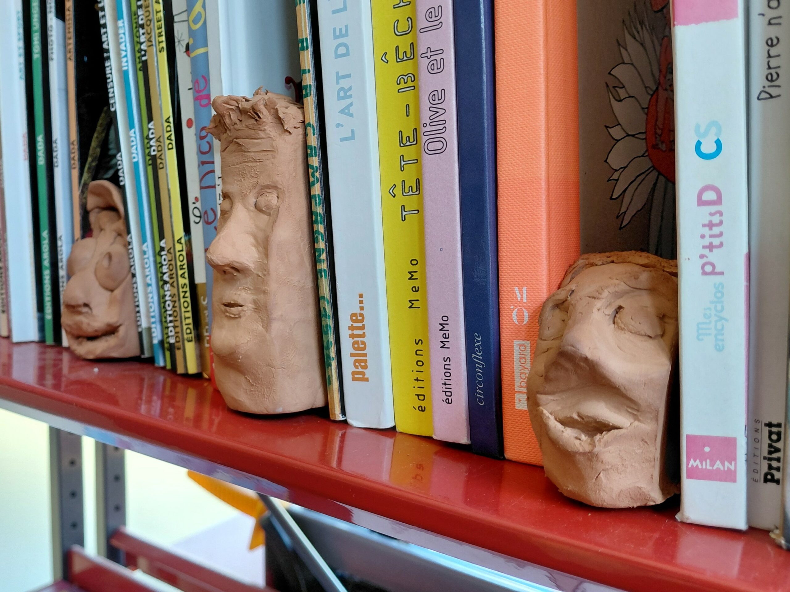 Visuel sur lequel on peut voir trois sculptures en argile glissés entre les livres d'une bibliothèque